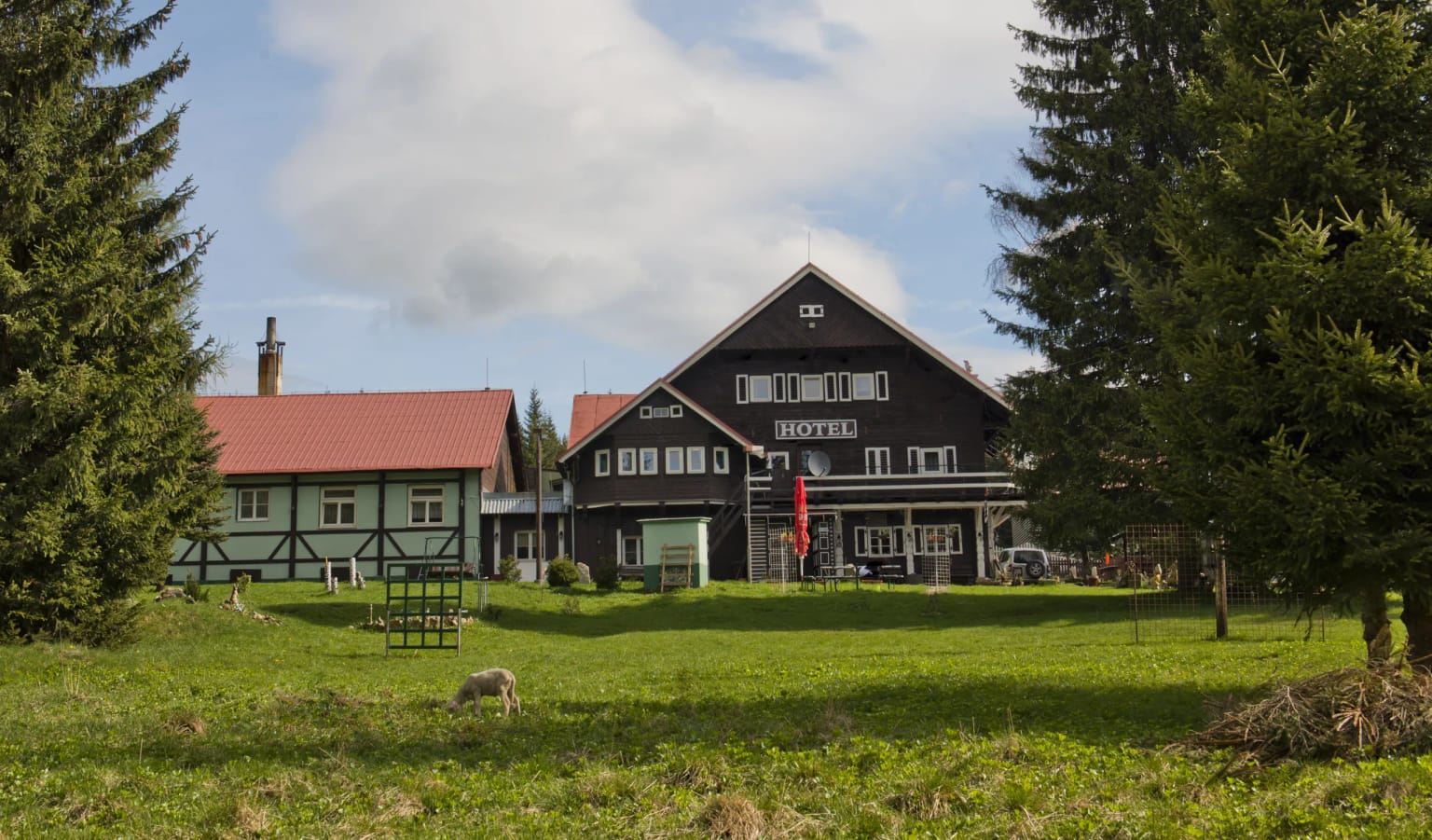 Hotel Červena Liška – dunkles Holzgebäude mit dem Schriftzug HOTEL und ein Nebengebäude mit rotem Dach auf einer grünen Wiese zwischen Fichten